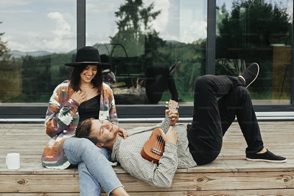 Stylish hipster couple sitting on wooden porch, relaxing with ukulele  on background of modern cabin with big windows in mountains. Happy young family travelers enjoying vacation in woods