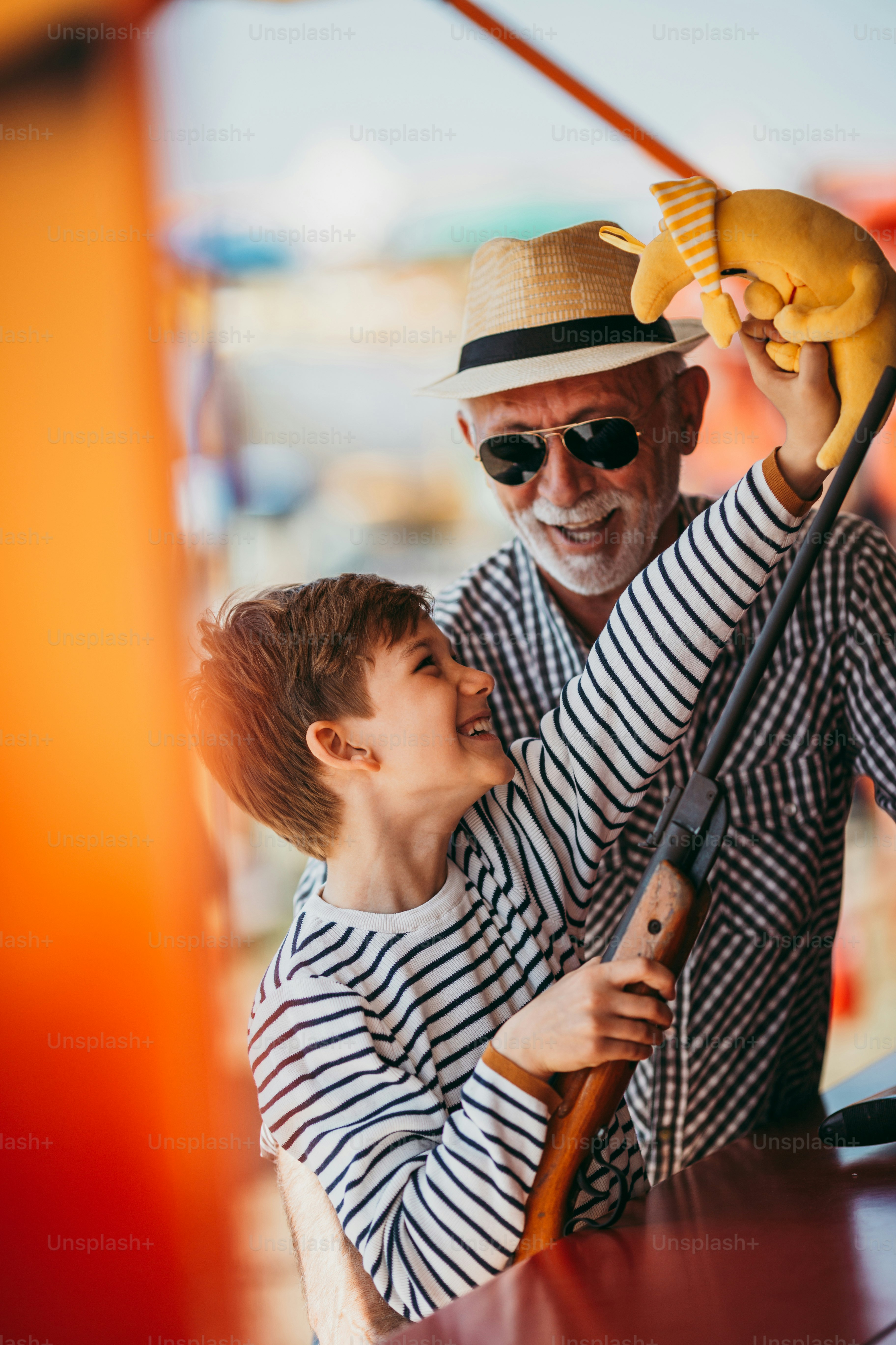 Abuelo y nieto divirtiéndose y pasando tiempo de buena calidad juntos en el parque de atracciones. Niño disparando con pistola de aire comprimido mientras el abuelo lo ayuda a ganar el premio.