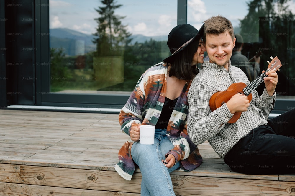 Happy hipster couple relaxing on wooden porch of modern cabin with big windows in mountains. Hipster man playing on ukulele for his stylish woman, enjoying romantic moment