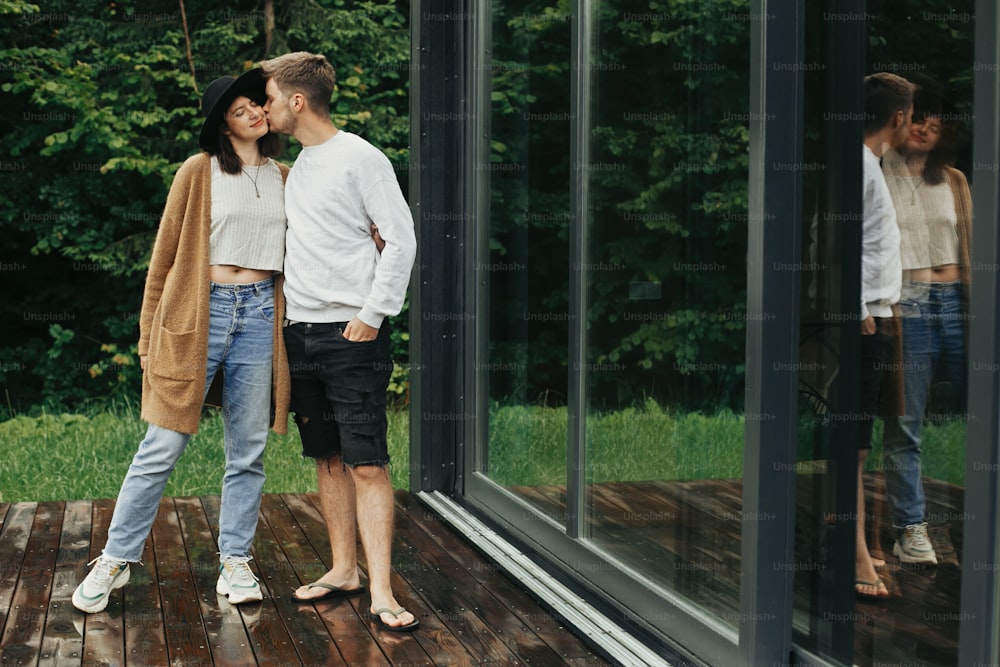 Stylish hipster couple hugging and kissing on wooden terrace, relaxing in cabin in mountains. Happy young family in modern outfits embracing on background of big window and trees