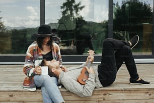 Hipster man playing on ukulele for his beautiful stylish woman, relaxing on wooden porch on background of modern cabin with big windows in mountains. Happy young family travelers enjoying vacation