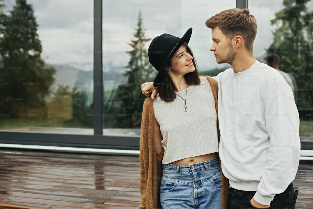 Stylish hipster couple hugging on wooden porch, relaxing in modern cabin in mountains. Happy young family in modern outfits embracing on background of terrace and big window