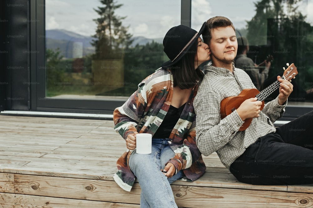 Happy hipster couple relaxing on wooden porch of modern cabin with big windows in mountains. Hipster man playing on ukulele for his stylish woman, enjoying new home in woods.