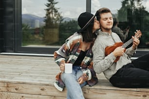 Feliz pareja hipster relajándose en el porche de madera de una cabaña moderna con grandes ventanales en las montañas. Hombre hipster tocando el ukelele para su elegante mujer, disfrutando de un nuevo hogar en el bosque.