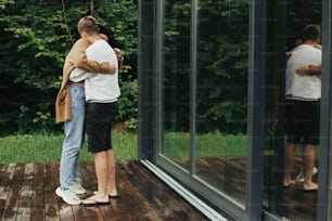 Elegante pareja hipster abrazando y besando en la terraza de madera, relajándose en una cabaña en las montañas. Familia joven feliz en trajes modernos que abrazan el fondo de la ventana grande y los árboles