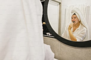Dentalhygiene. Null Abfall. Schöne junge Frau in weißem Handtuch putzt ihre Zähne mit Bambuszahnbürste und Kohlezahnpasta im modernen Badezimmer, Blick auf runden Spiegel.