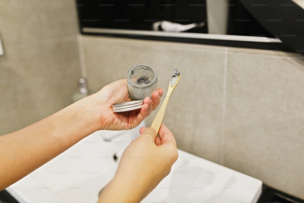 Woman using bamboo toothbrush and charcoal toothpaste from glass jar for daily cleaning routine. Recycle and reuse. Wooden toothbrush with paste closeup. Zero waste bathroom concept