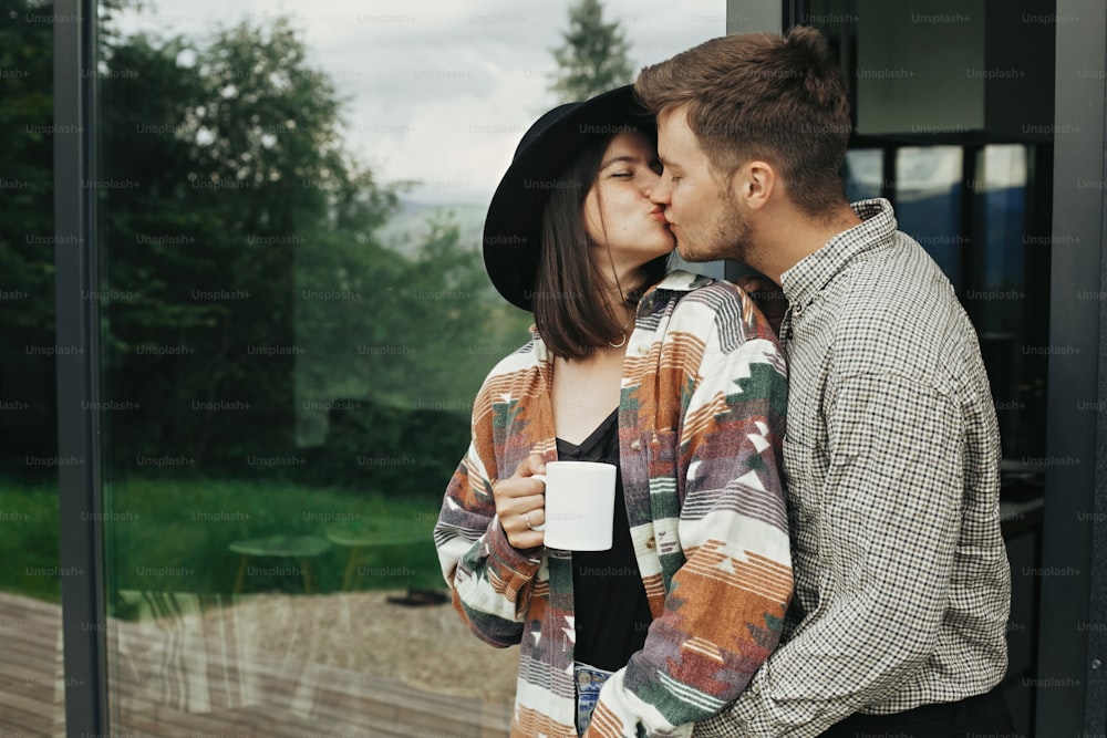 Elegante pareja hipster con café de la mañana besándose en el fondo de una cabaña moderna con grandes ventanas en las montañas. Familia joven feliz abrazando y disfrutando de un nuevo hogar en el bosque. Espacio para el texto