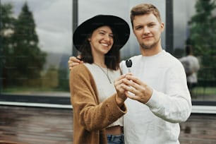 Home and family concept. Stylish hipster couple holding together key from their new home on background of terrace and big windows outdoors. Happy young family of homeowners, purchasing real estate