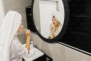 Beautiful young woman in white towel brushing her teeth with bamboo toothbrush and charcoal toothpaste in modern bathroom, looking at round mirror. Dental hygiene. Zero waste.