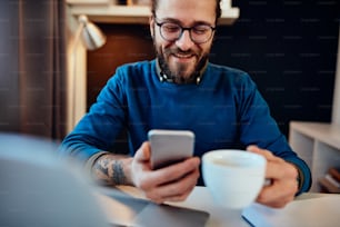 Good-looking caucasian young businessman sitting in his office, holding cup of coffee and looking at smart phone. Selective focus on smart phone.