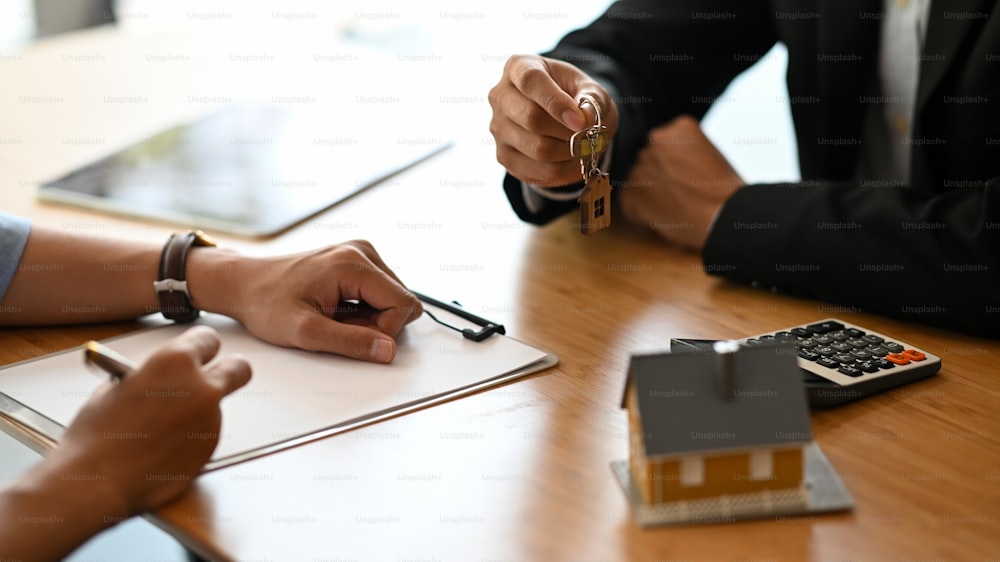 Abgeschnittene Aufnahme des Hausmaklers, der seinem Kunden einen Immobilienschlüssel gibt, während er am modernen Holzschreibtisch schreibt / unterschreibt. Kredit-, Schulden-, Kredit-, Kauf- oder Verkaufsvertragskonzept.