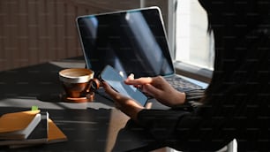 Side shot of young businesswoman holding white blank screen smartphone in her hand while sitting at the modern working desk with laptop, notebook and coffee cup as background.