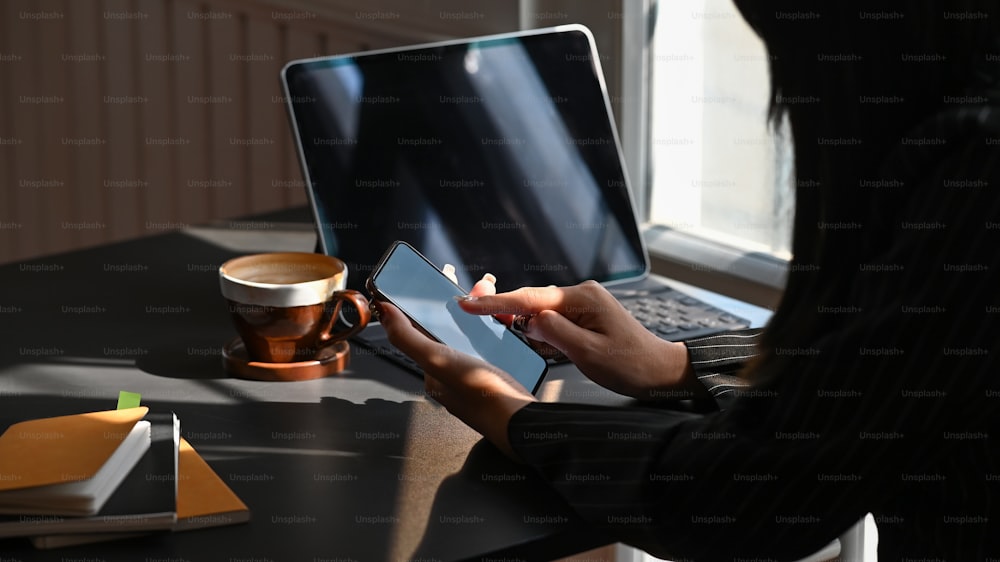 Side shot of young businesswoman holding white blank screen smartphone in her hand while sitting at the modern working desk with laptop, notebook and coffee cup as background.