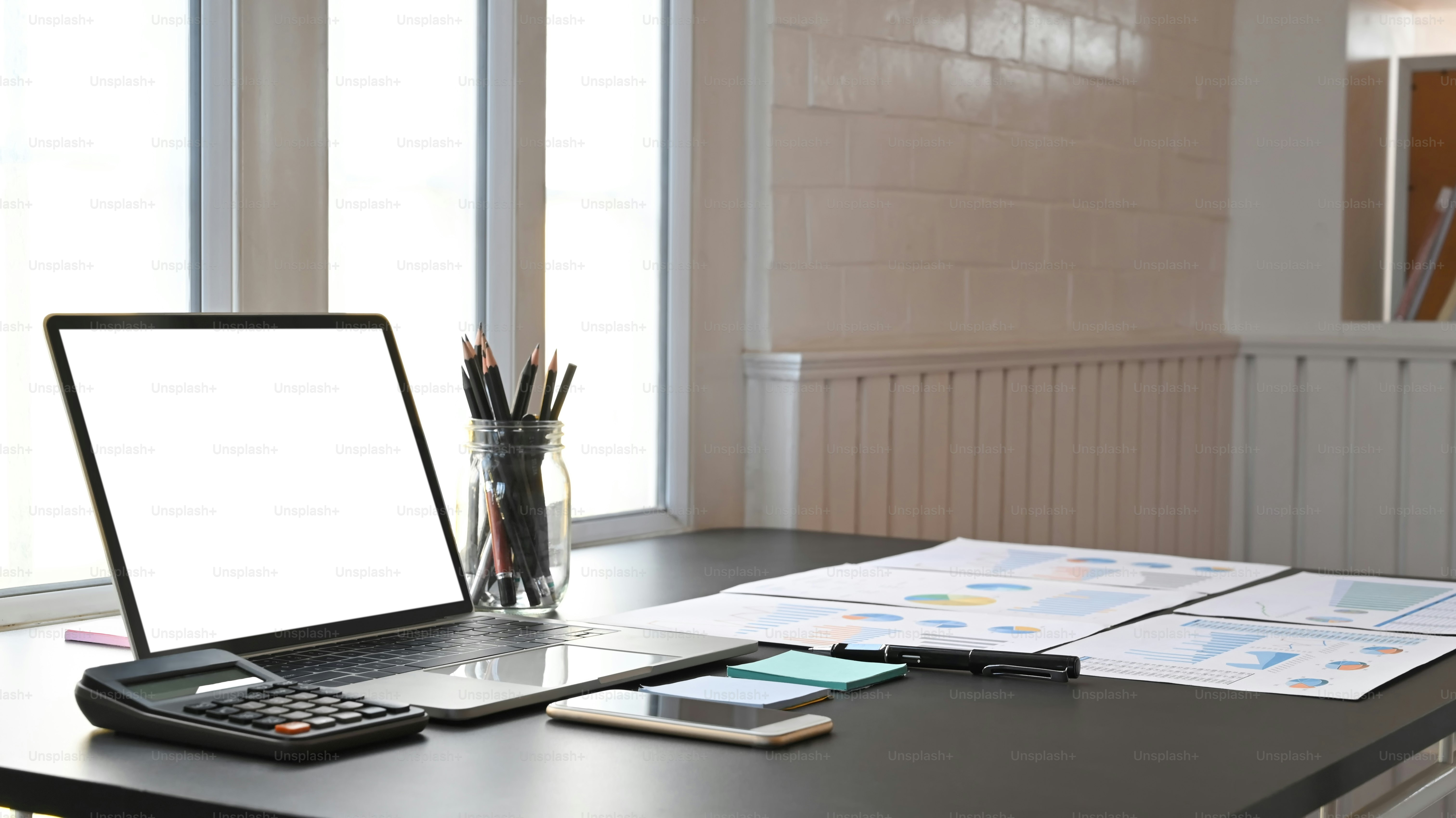 Photo of business equipment/accessories putting on the modern working desk. Flat lay white blank screen laptop, smartphone, calculator, pencil holder, post-it and graphic chart on the black table.