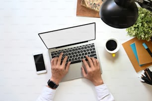 현대적인 작업 책상에 흰색 빈 화면 노트북에 타이핑하는 창의적인 남자의 상위 뷰. 검은색 빈 디스플레이, 커피잔, 노트북, 램프, 연필꽂이, 화분에 심은 스마트폰.