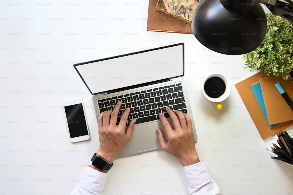 Vista superior del hombre creativo que escribe en una computadora portátil con pantalla blanca en blanco en el escritorio de trabajo moderno. Teléfono inteligente con pantalla vacía negra, taza de café, cuaderno, lámpara, portalápices y planta en maceta.
