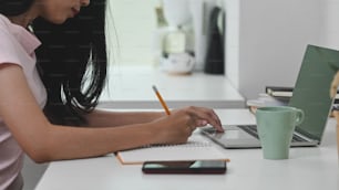 Cropped shot of young beautiful woman writing/taking notes while sitting at the modern working desk with comfortable living room as background.