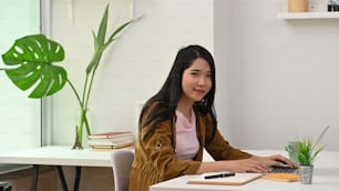 Young creative woman smiling and looking at the camera while sitting at the modern working desk with comfortable office as background.