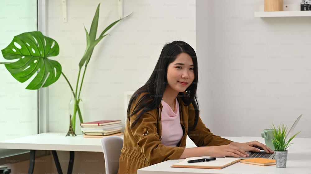 Young creative woman smiling and looking at the camera while sitting at the modern working desk with comfortable office as background.