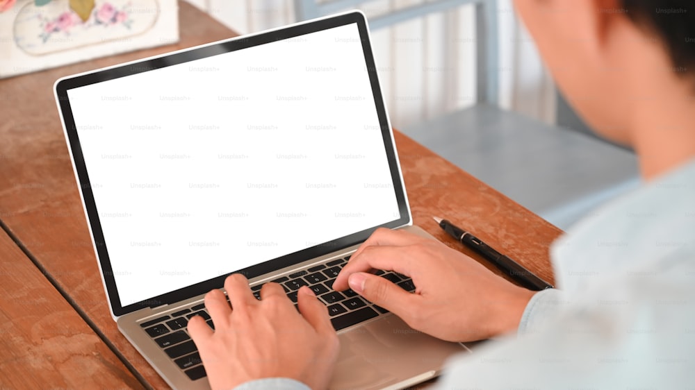 현대적인 나무 책상에 앉아 흰색 빈 스크린 노트북에 타이핑하는 창의적인 회사원의 사진.