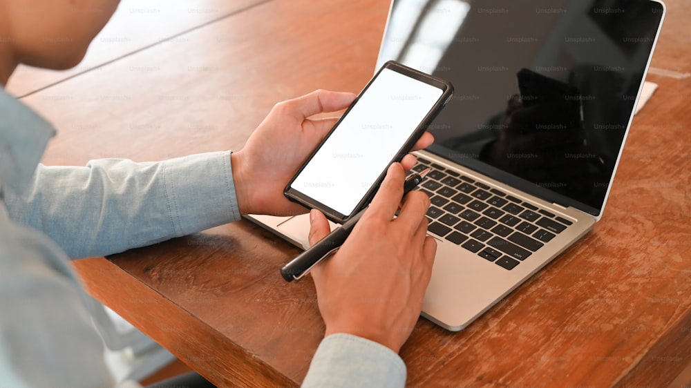 Foto cortada de um empresário segurando um smartphone branco em branco e uma caneta na mão enquanto estava sentado em frente ao laptop de tela preta na mesa de trabalho de madeira.