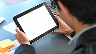 Abgeschnittene Aufnahme eines smarten Geschäftsmannes im schwarzen Anzug, der ein schwarzes Tablet mit weißem leerem Bildschirm in der Hand hält, während er am modernen Tisch mit modernem Besprechungsraum als Hintergrund sitzt.