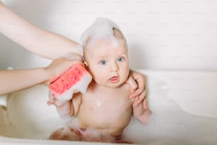Happy laughing baby taking a bath playing with foam bubbles. Little child in a bathtub. Infant washing and bathing. Unrecognizable mother bathing her baby in white small plastic bat