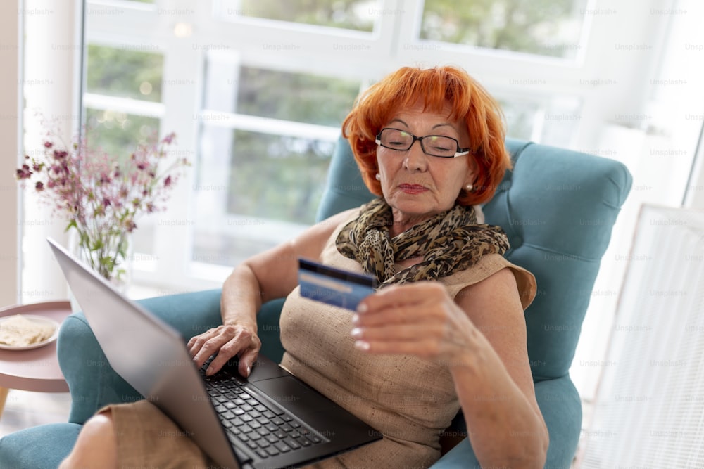 Beautiful elderly woman sitting in an armchair, relaxing at home, using a laptop computer, paying online using a credit card; senior woman doing Internet shopping