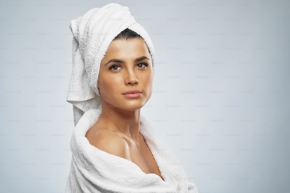 Side view of pretty female with towel on head and in bathrobe posing. Portrait of brunette woman with naked shoulder looking at camera, isolated on grey background. Beauty, skincare concept.