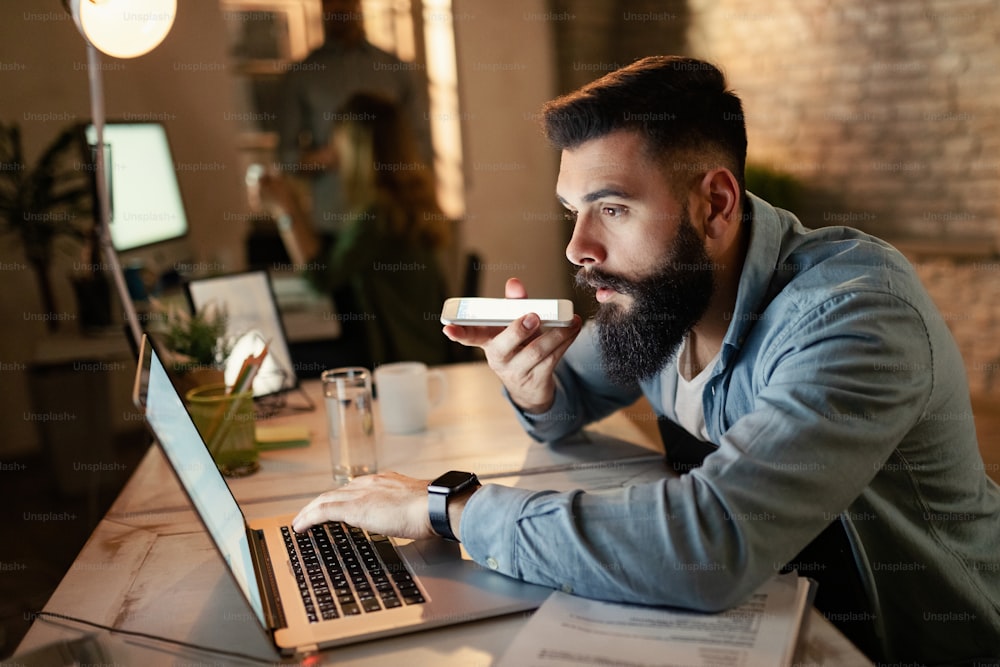 Jeune entrepreneur lisant un e-mail sur un ordinateur tout en enregistrant un message vocal sur un téléphone intelligent pendant le travail de nuit au bureau.