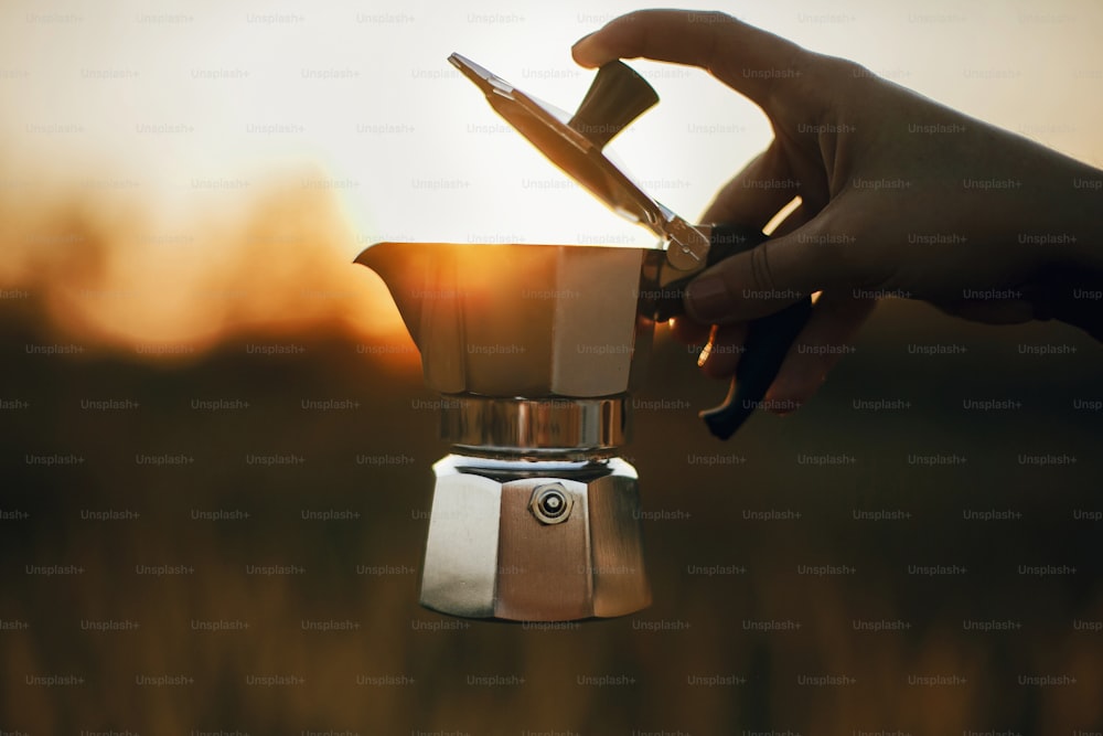 Viaggiatore che tiene la caffettiera Geyser con caffè fresco in luce calda e soleggiata nelle erbe della campagna rurale. Momento rustico atmosferico. Preparazione alternativa del caffè in viaggio.