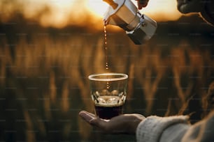 Preparación alternativa de café en viajes. Verter café caliente recién hecho de la cafetera del géiser en una taza de vidrio con una luz cálida y soleada en hierbas rurales del campo. Momento atmosférico. Imagen vertical