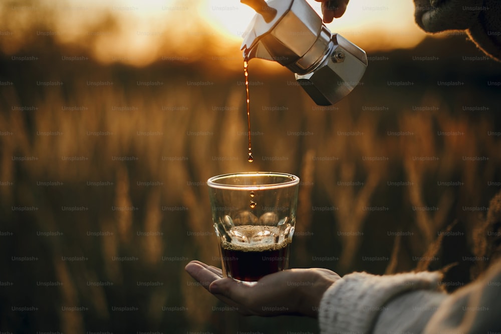 Alternative coffee brewing in travel. Pouring fresh hot coffee from geyser coffee maker into glass cup in sunny warm light in rural countryside herbs. Atmospheric moment. Vertical image