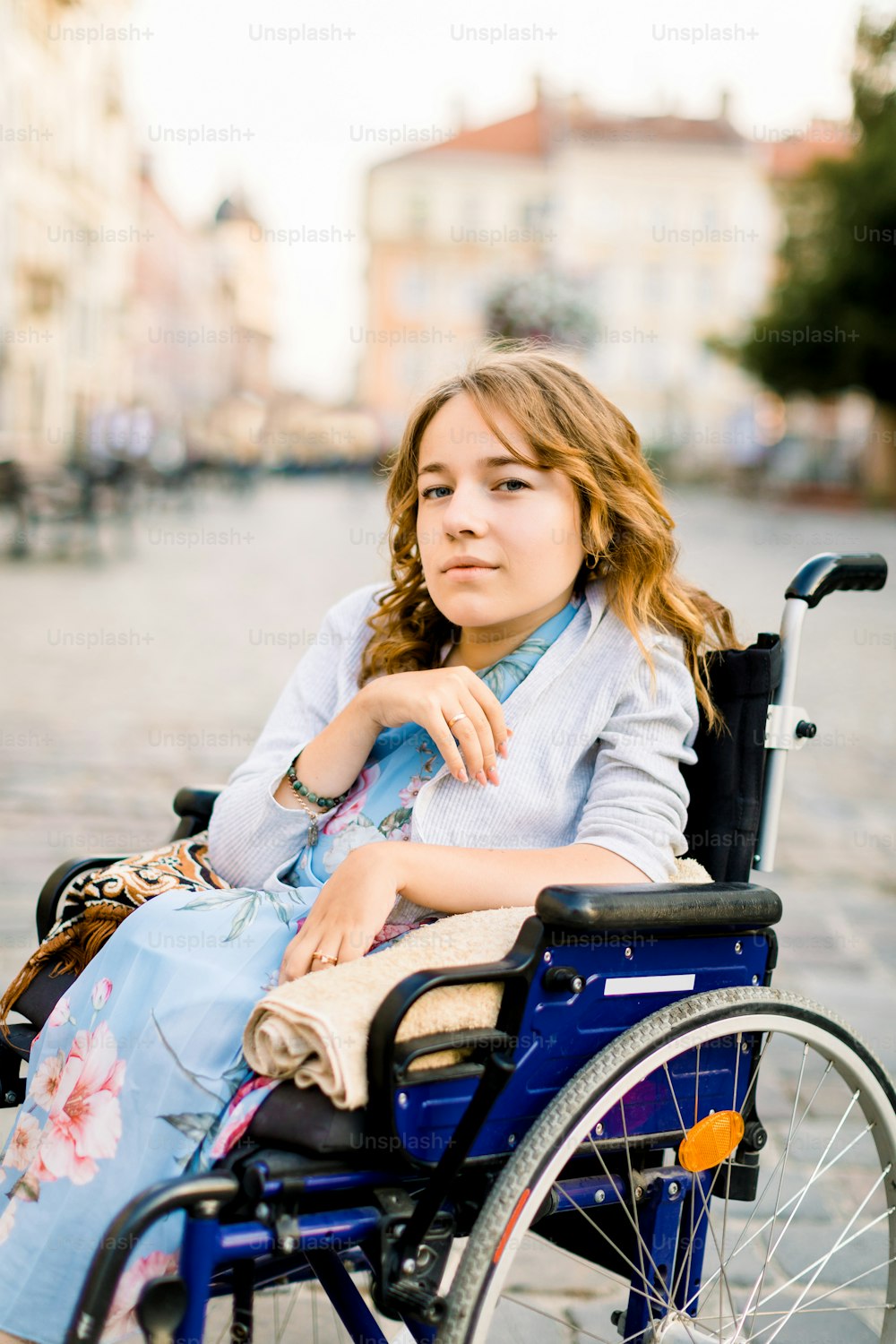 Portrait of young blond-haired woman sitting in wheelchair and looking at camera, walking outdoors in old city center.