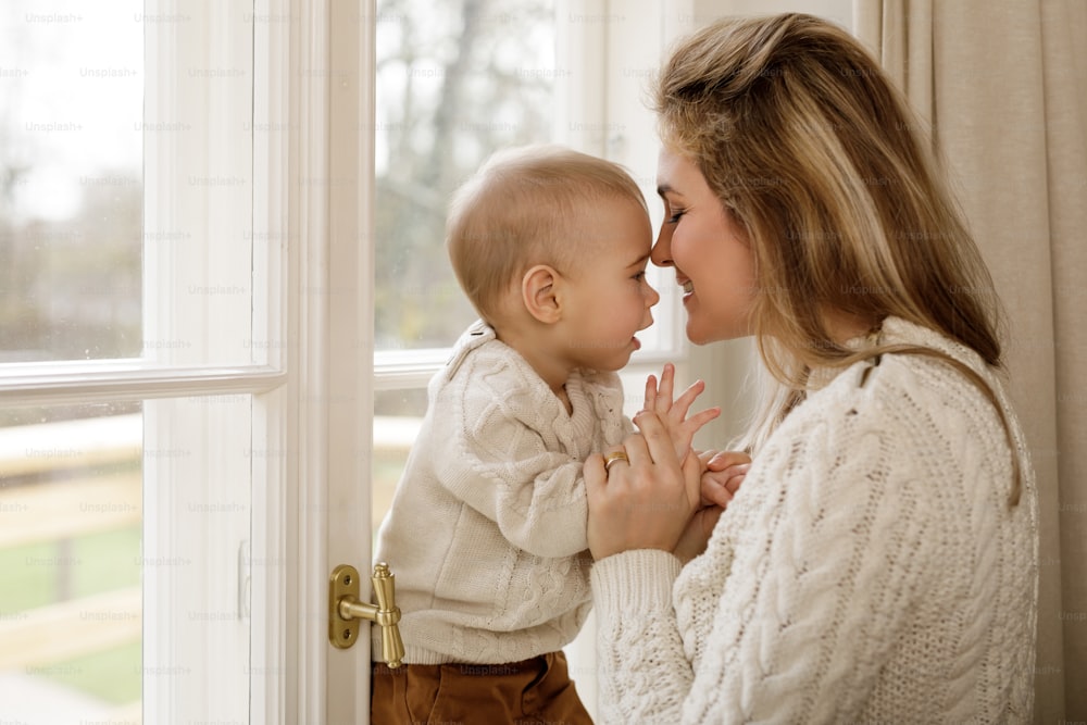 Young and beautiful mother and her little baby son wearing warm sweaters standing by the window