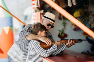 Grandfather and grandson having fun and spending good quality time together in amusement park. Kid shooting with air gun while grandpa helps him to win the prize.