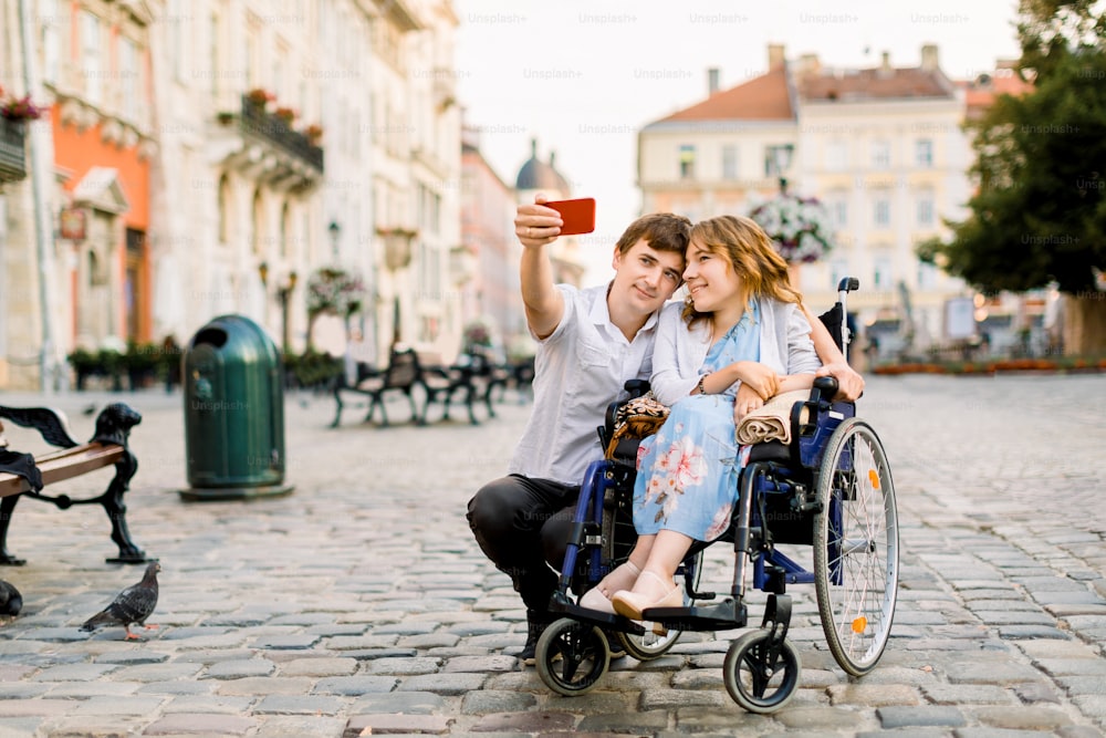 Selfie time. Handsome happy young man hugging his disabled young pretty woman and smiling while taking selfie with her in the city.