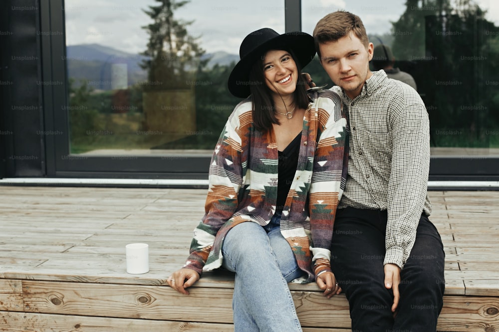 Stylish hipster couple with morning coffee sitting on wooden porch, relaxing on background of modern cabin with big windows in mountains. Happy young family travelers enjoying vacation in woods