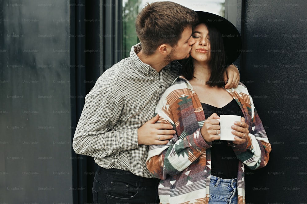 Stylish hipster couple with morning coffee relaxing on background of modern cabin and big windows in mountains. Happy young family enjoying morning at new home in woods. Travel and vacation