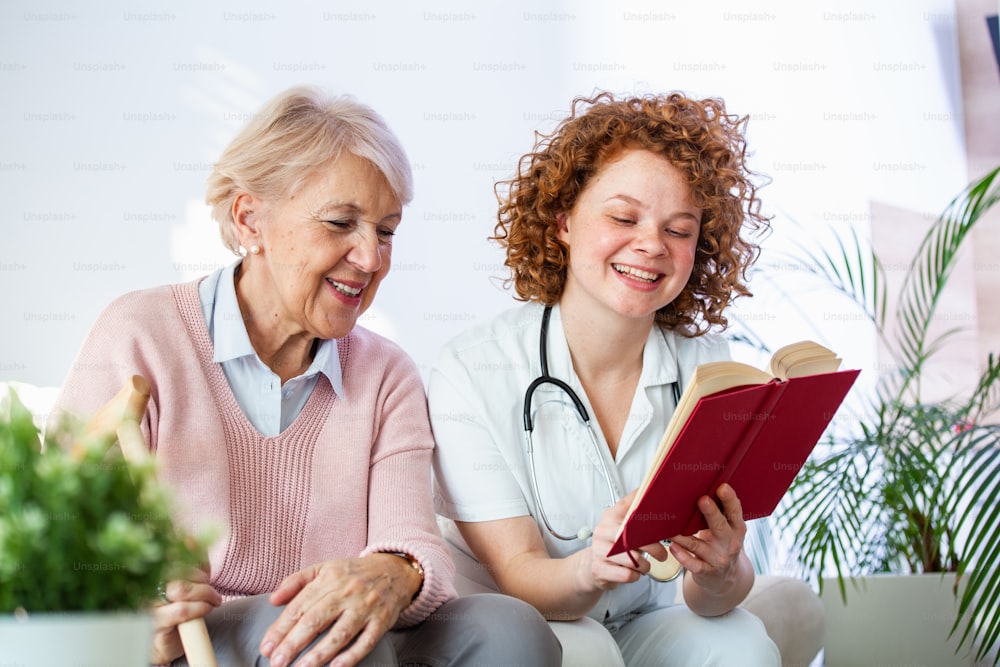 Woman caregiver reading a book while sitting with happy senior woman at nursing home. Happy elder woman sitting on white sofa and listening to nurse reading a book out loud
