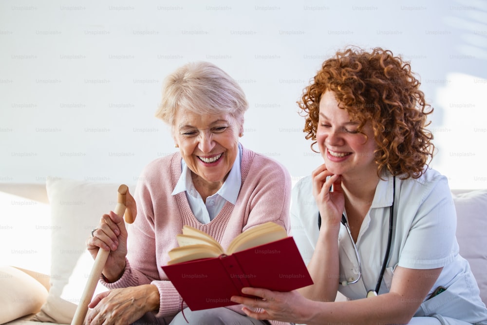Cuidadora leyendo un libro mientras está sentada con una mujer mayor feliz en un hogar de ancianos. Anciana feliz sentada en un sofá blanco y escuchando a la enfermera leyendo un libro en voz alta