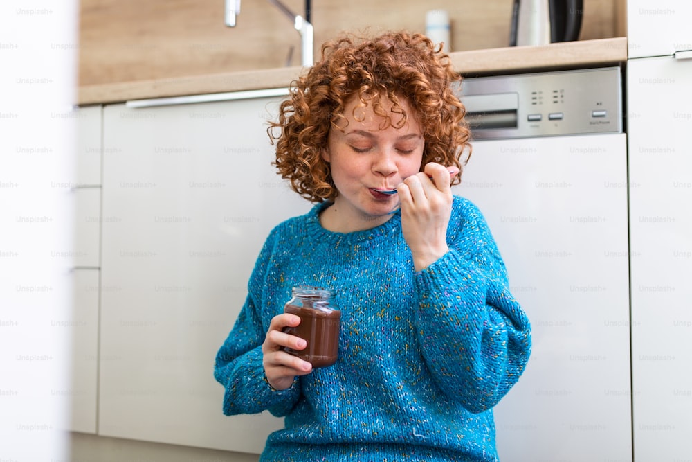 young woman eating chocolate from a jar while sitting on the wooden kitchen floor. Cute ginger girl indulging cheeky face eating chocolate spread from jar using spoon savoring every mouthful