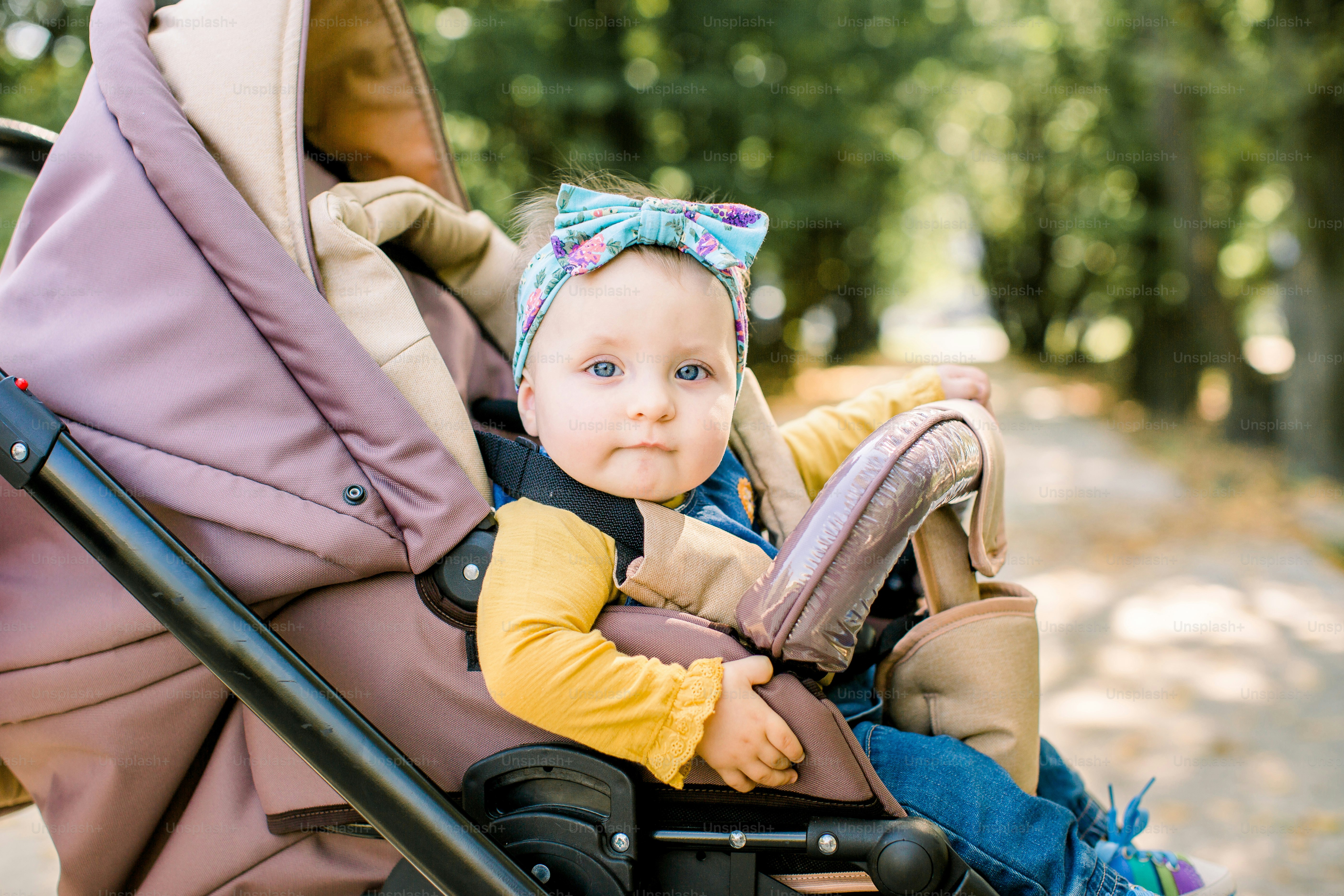 Happy girl in baby carriage playing in pram on background nature. Portrait cute little beautiful girl of 9 months sitting or stroller and waiting for mom