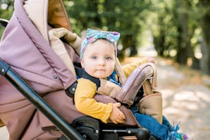 Happy girl in baby carriage playing in pram on background nature. Portrait cute little beautiful girl of 9 months sitting or stroller and waiting for mom