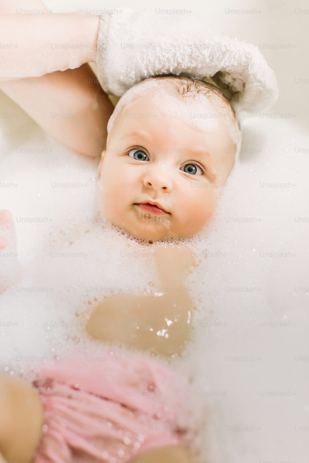 거품 거품을 가지고 노는 목욕을 하는 행복한 웃는 아기. 욕조에 있는 어린 아이. 유아 씻기 및 목욕. 어린 아이들을위한 위생 및 보살핌. 머리에 비누 거품을 가진 사랑스러운 목욕 아기