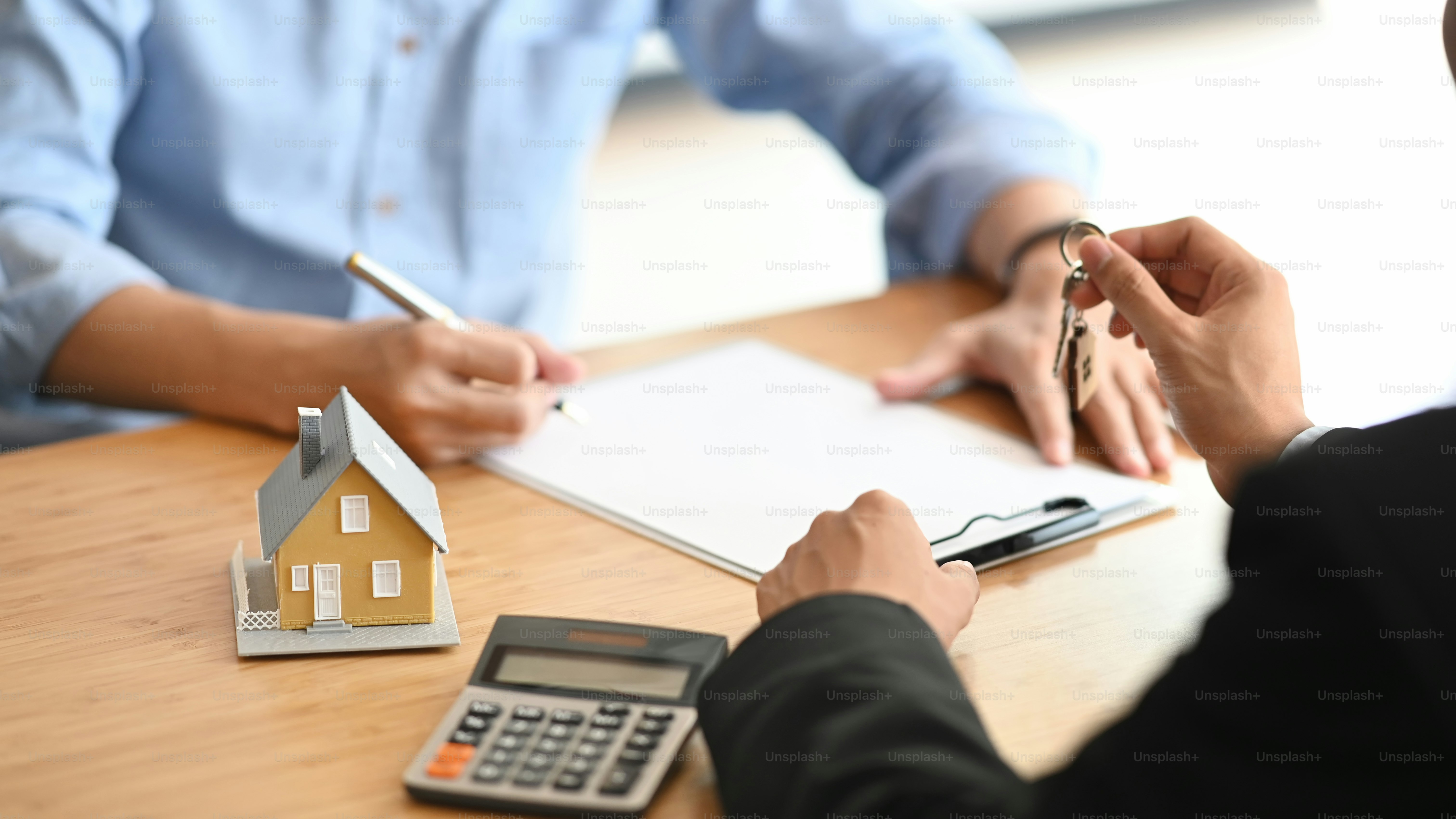 A person holding keys near a model house and calculator on a desk