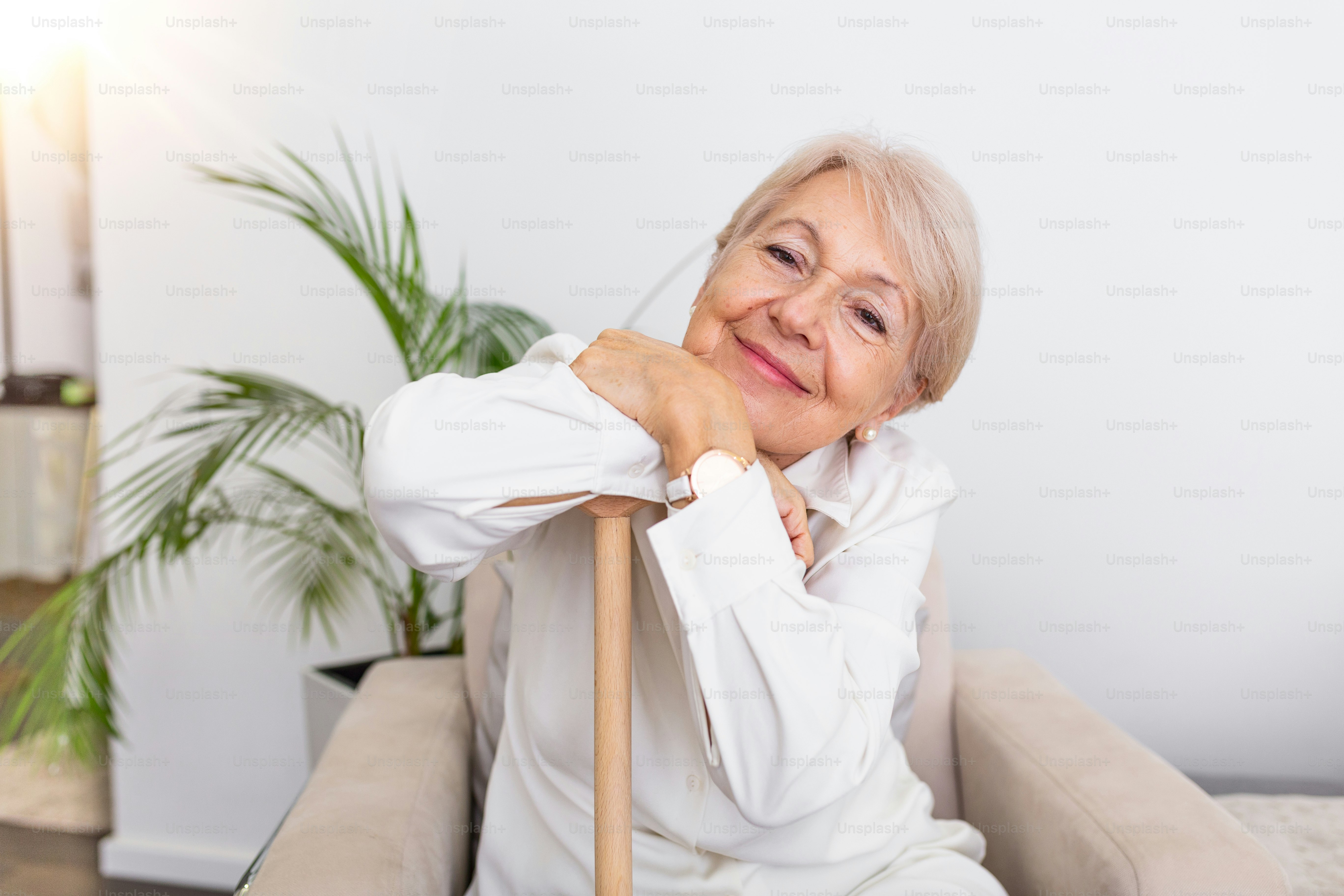 Smiling grandmother sitting on couch. Portrait of a beautiful smiling senior woman with walking cane on light background at home. Old woman sitting with her hands on a cane