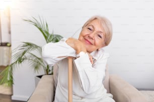 ソファに座る笑顔のおばあちゃん。自宅で明��るい背景に杖をついて歩く美しい笑顔の年配の女性の肖像画。杖に手をついて座っている老婆