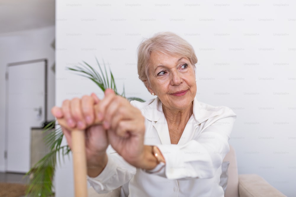 Elder lady sitting on the couch with wooden walking stick and smiling. Happy elderly woman relaxing on sofa and holding walking stick. Copy space. Senior woman looking thoughtful in a retirement home